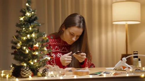 A Woman Dressed in a Festive Sweater Works on a Craft Project, Surrounded by Christmas Decorations a