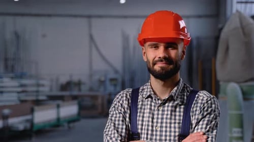 Porträt eines bärtigen Fabrikarbeiters mit Schutzhelm, der in die Kamera blickt, während er in der Werkstatt steht