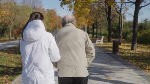 Elderly Man with Supportive Nurse Back View of Autumn Park Stroll