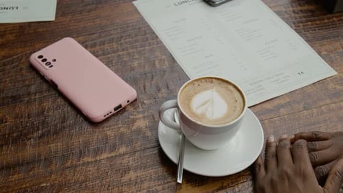 Hands of Unknown Woman Sitting at Table Featuring Phone, Menu and Cup of Coffee