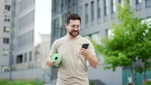 A happy male sportsman with a mat in his hands is walking along city street on a training looking