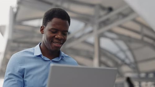 Un homme afro-américain souriant travaillant à distance sur l'ordinateur portable de l'extérieur, beau multiethnique