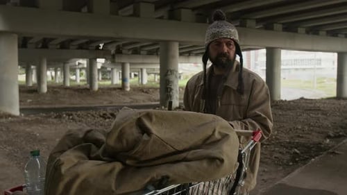 Homeless Man Looking around and Pushing Cart with Belongings under Bridge