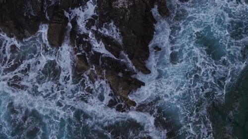 Drone view of turbulent ocean waves surging around dark coastal rocks.