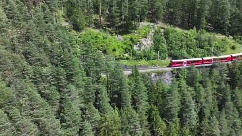 Drone rotates to track red railway train passing to tunnel of majestic green mountains and emerging