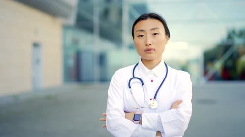 Close up portrait of Asian woman doctor looking at camera with arms crossed on background of modern