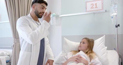 Diverse male doctor showing girl patient in hospital bed how to use asthma inhaler, slow motion