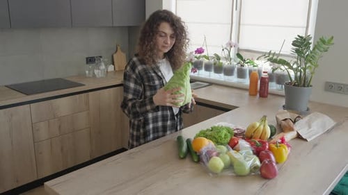 Smiling Woman Holding Lettuce And Admiring Fresh Groceries In Sunlit Kitchen