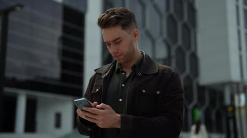 Portrait of Focused Handsome Young Man Using Typing Smartphone Standing Outside Modern Office Center