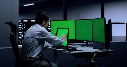 Programmer Looking At Green Screen Tablet While Working On Green Screen Desktops In The Server Room