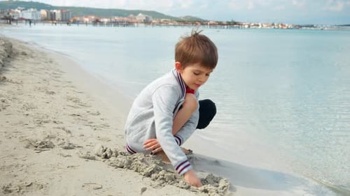 A young boy sits on a sunny beach digging with hands warm sand