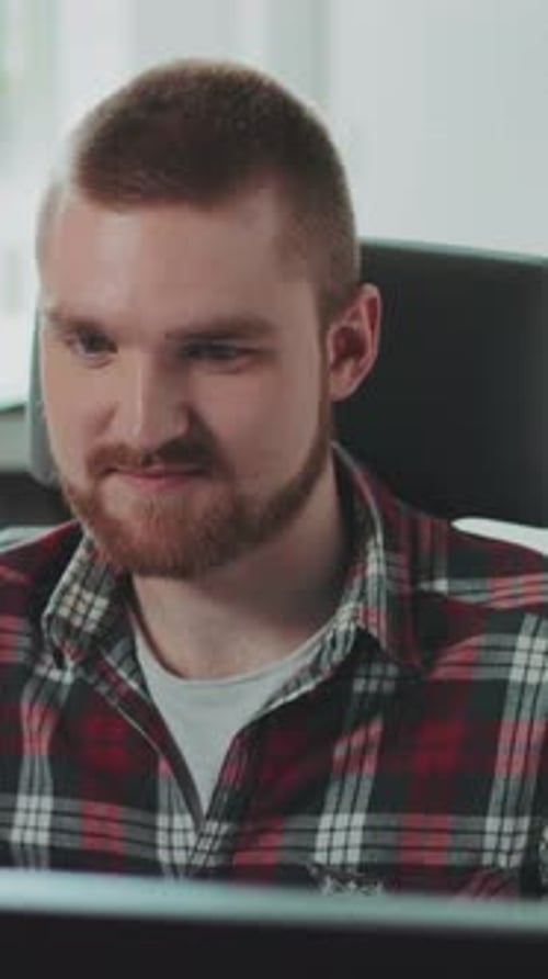 Ein junger bärtiger Mann sitzt an einem Computer, arbeitet im Büro, fühlt sich glücklich, zeitgenössisches Design, gedreht