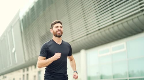 Young Caucasian bearded happy man runner jogging down the street on urban background of city park.