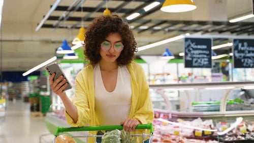 Focused Young Woman in Supermarket Uses Smartphone to Check Purchased Groceries