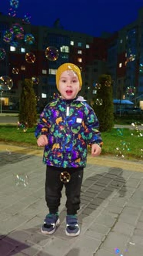 Happy energetic kid in warm clothes jumping cheerfully among the soap bubbles.