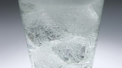 Close up of ice in a glass into which a fizzy clear drink is slowly poured. Cool refreshment with mi