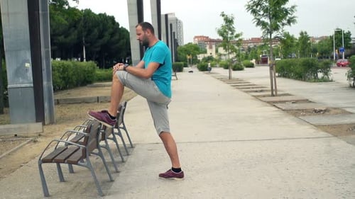 Young man stretching legs on bench in city park in super slow motion