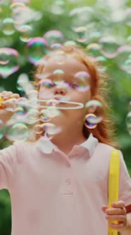 Young Girl Blows Bubbles in a Park Surrounded By Greenery on a Sunny Day