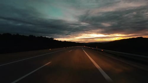 Driving on empty long asphalt road in the night with dusk sunset in background over a cloudy sky