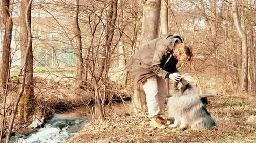On this video you see a flowing brook, a young blond man with medium length hair petting his dog, a