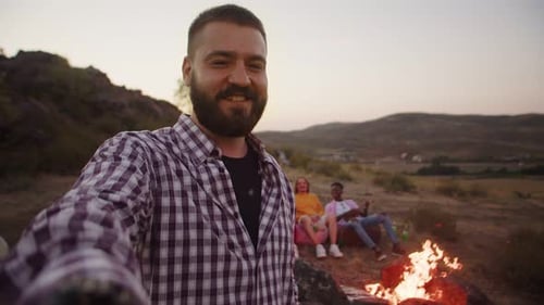 Young Handsome Bearded Caucasian Man Shooting Selfie Video About His Friends Sitting in the Evening