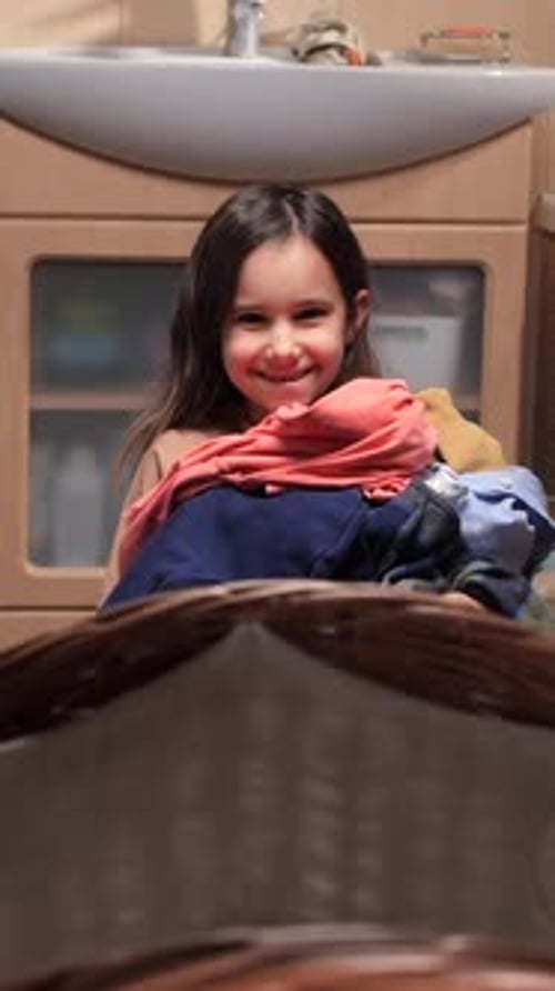Vertical Video Happy Young Girl Holding a Pile of Colorful Clothes Above a Wicker Laundry Basket