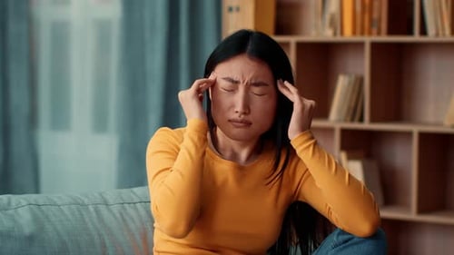 Woman With Long Dark Hair Sitting on Couch Holding Head in Pain at Home