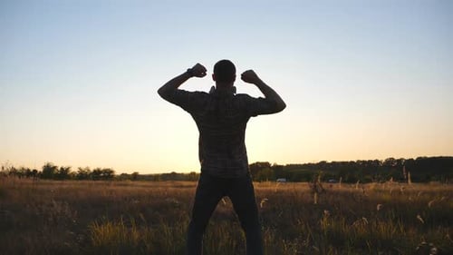 Young Man Victoriously Raising Hands Up on Grass Field at Sunset