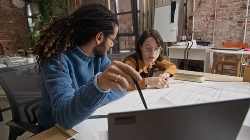Two Diverse Colleagues Collaborating on Laptop with Floor Plan at Desk in Office