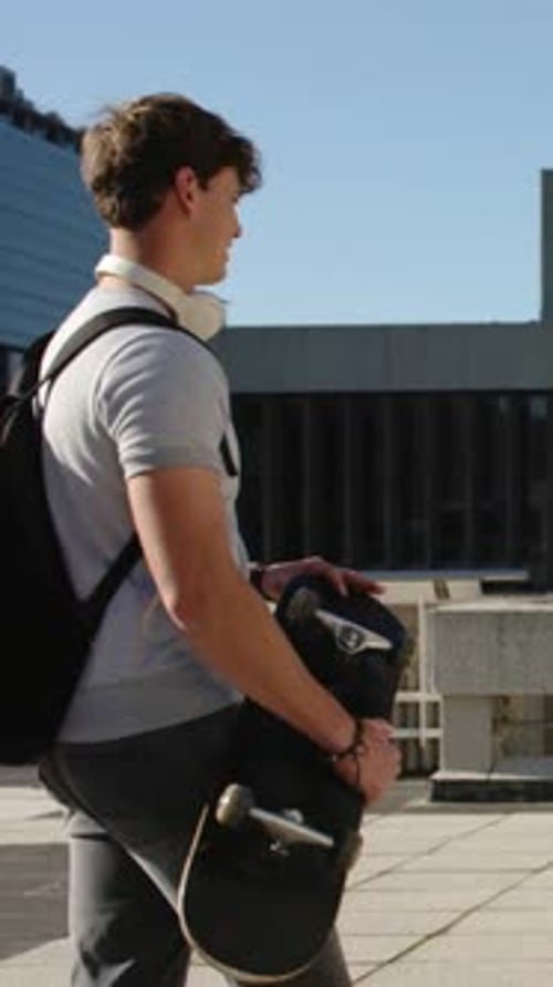 Vertical video: Entering left man carrying skateboard walking plaza past low barrier, headphones