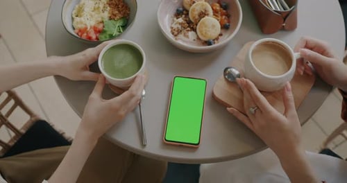Closeup of Female Hands Holding Coffee Cups Next to Smartphone with Green Screen Chroma Key at Table