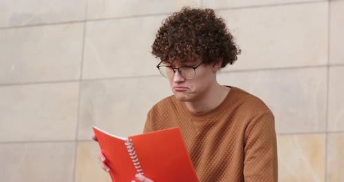 Man opening orange notebook, reading while frowning, tilting it, flipping pages on courtyard bench