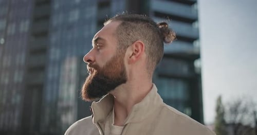 A Young Man with Stylish Trendy Hairstyle and Beard in Casual Clothes Stands on the Street in the