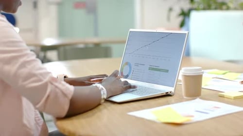 Afroamerikanisches Team arbeitet am Bürotisch, als ein Kollege Kaffee trinkt und Laptop-Charts überprüft