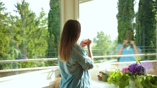 Joyful Caucasian Beautiful Woman in Positive Mood Standing Near Window with Cup of Coffee or Tea in
