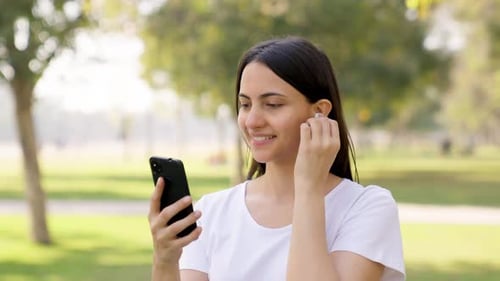 Woman Inserts Wireless Earbud While Using Phone Outdoors