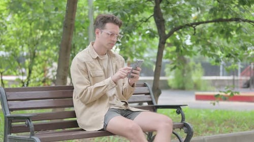 Man Uses Smartphone Sitting on Park Bench