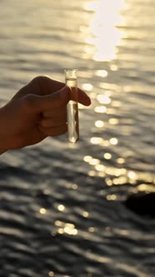 Hand holding a test tube filled with water, taking a sample from a lake or sea during sunset