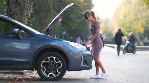 Puzzled Female Driver Standing on a Street Near Her Car with Popped Up Hood Inspecting Broken Engine