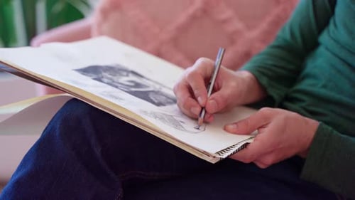 Close up of arstist's hands drawing woman's face on sketch pad while sitting on pink couch
