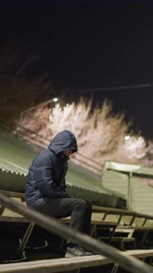 Man in Hooded Jacket Sitting Alone on Bleachers at Night with Distant Car
