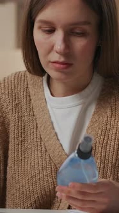Mujer joven pensativa con un acogedor cárdigan de punto que examina la botella de aerosol y luego trabaja concentrada en el escritorio