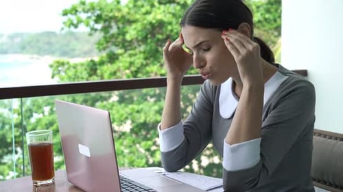 Woman with headache using laptop on tropical balcony