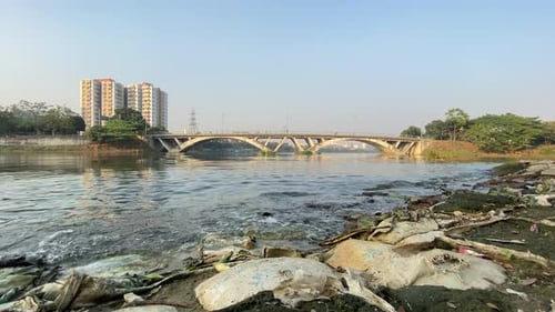 Riverbank View Beside Rubbish Of Tidal Waves With Bridge With Traffic Going Past. Low Angle, Locked