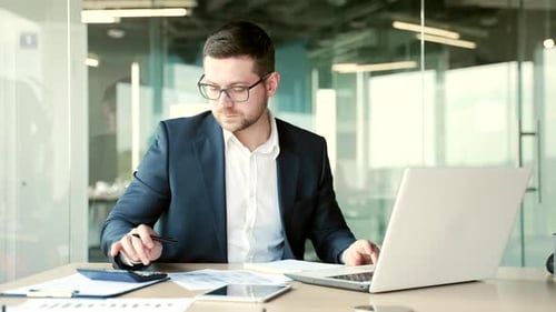 Busy financier in formal suit making financial calculations using calculator, typing on a laptop