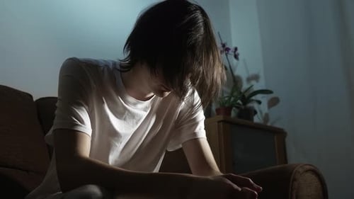 Jeune homme assis pensivement dans la solitude dans une pièce sombre, ressentant une lourdeur émotionnelle, personne penchée