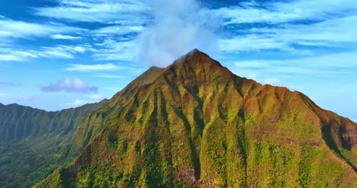Approaching a sharp peak of the verdant mountain. Azure sky with light clouds at backdrop.