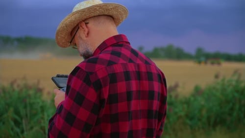 Man Farmer in Straw Hat Controls Harvesting Process Using Drone for Observation Combine Harvester in