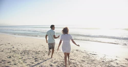Biracial couple runs along a sunlit beach with copy space, their backs to the camera