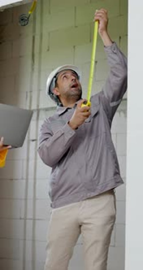Asian male contractor measures vertical wall with tape while woman engineer records laptop data,